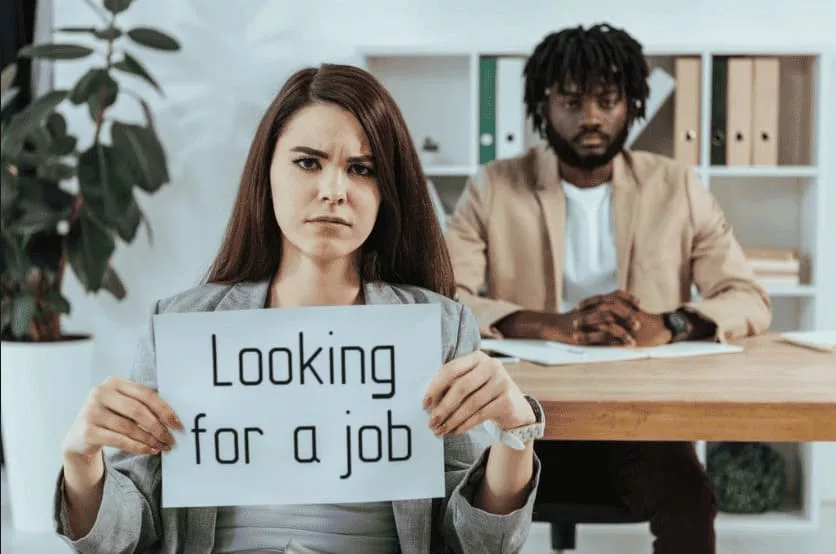Mujer con expresión seria sosteniendo un cartel que dice "Looking for a job" en una entrevista de trabajo, con un hombre sentado al fondo. onhunters, firma de búsqueda y selección de personal.
