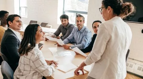 Líder femenina dirigiéndose a un grupo diverso de profesionales en una reunión de equipo colaborativa.