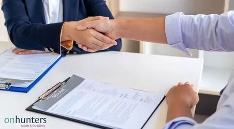 Candidato y reclutador estrechando la mano tras una entrevista de trabajo, con un currículum sobre la mesa. onhunters, firma de búsqueda y selección de personal.