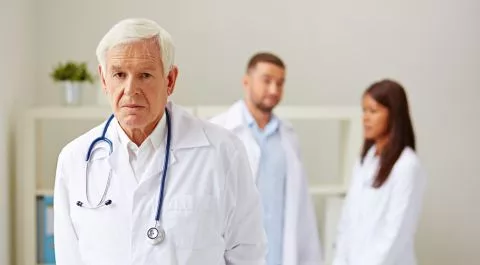 Médico senior de cabello blanco con bata blanca en primer plano, con dos profesionales jóvenes desenfocados al fondo.
