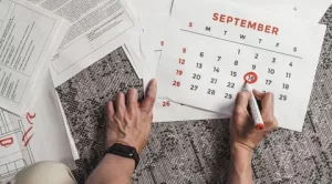 Manos marcando el 16 de septiembre en un calendario de pared, con documentos de oficina esparcidos en el escritorio.