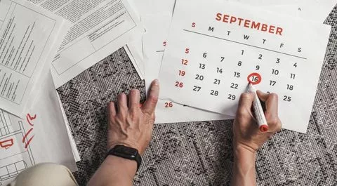 Manos marcando el 16 de septiembre en un calendario de pared, con documentos de oficina esparcidos en el escritorio.
