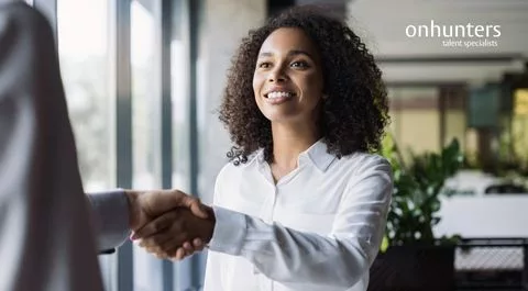 Dos personas, una mujer sonriente y un hombre, se dan la mano en una oficina, simbolizando una conexión profesional.
